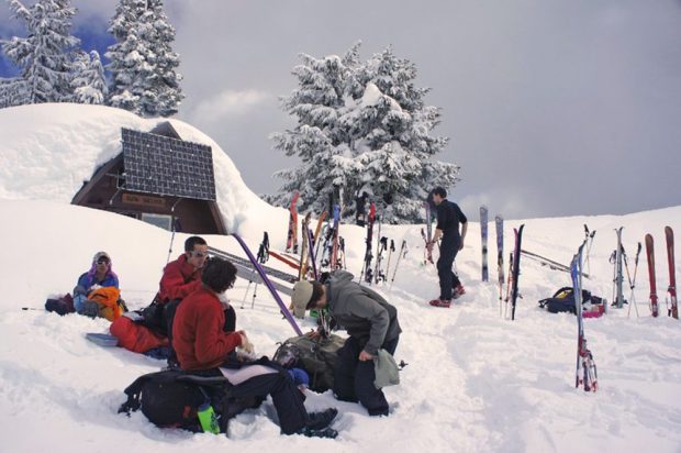 Trois hommes assis sur leurs sacs à dos à l’abri de la neige profonde prennent un casse-croûte devant le refuge. Les skis et les bâtons de ski sont coincés dans la neige se trouvant derrière le groupe se sustentant et un autre membre s’approche de l’entrée du refuge.