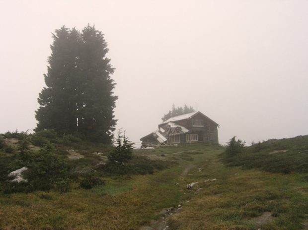 Photo du gîte prise après de nombreuses années de désuétude, avec le toit écroulé sur le côté gauche. Deux grands arbres à feuilles persistantes se dressent à l’extrémité gauche du gîte, et le sentier menant à ce dernier est couvert d’herbes et de petits arbustes.