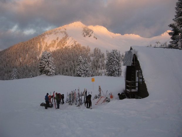 Un groupe de skieurs se tenant près du matériel de ski coincé dans la neige près de l’entrée principale d’un refuge. Le refuge est recouvert d’une couche de neige et les rayons du soleil transpercent de sombres nuages gris au loin.