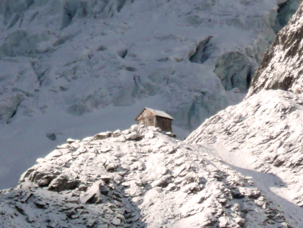 Un abri de pierres doté d’une chambre en A occupant la cime d’une crête montagneuse enneigée et étant baigné de soleil. Un long versant recouvert de neige est visible derrière l’abri en arrière-plan.