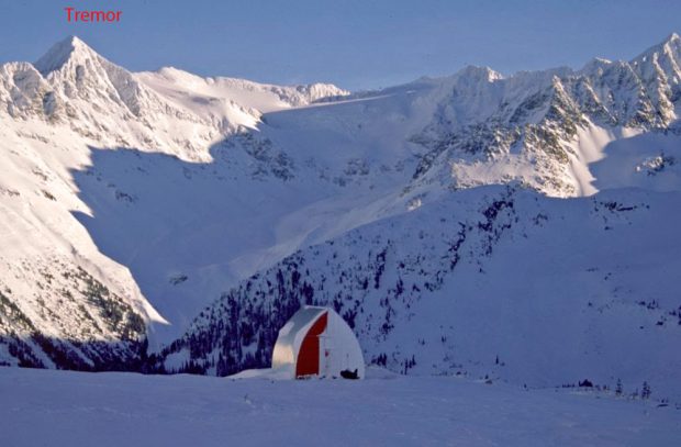 Une partie du refuge Himmelsbach est libre de neige, ce qui découvre le mur d’extrémité rouge et le revêtement en aluminium du toit. Le soleil brille sur les sommets enneigés en arrière-plan.