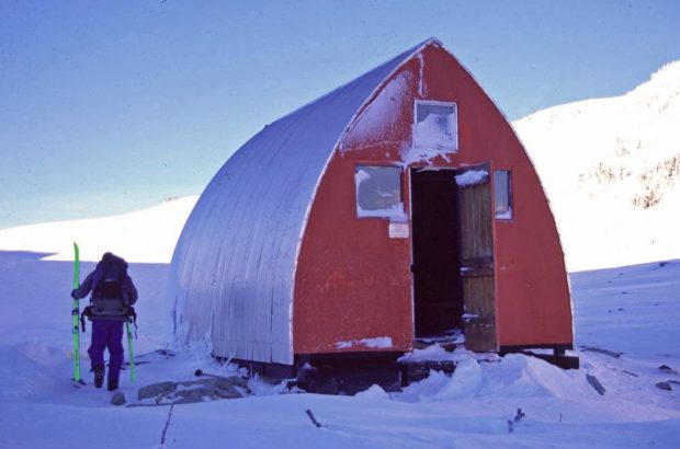 Un homme tenant des skis de randonnée vert fluo contourne le refuge, la porte avant est entrebâillée et le soleil brille sur les pentes abruptes situées derrière