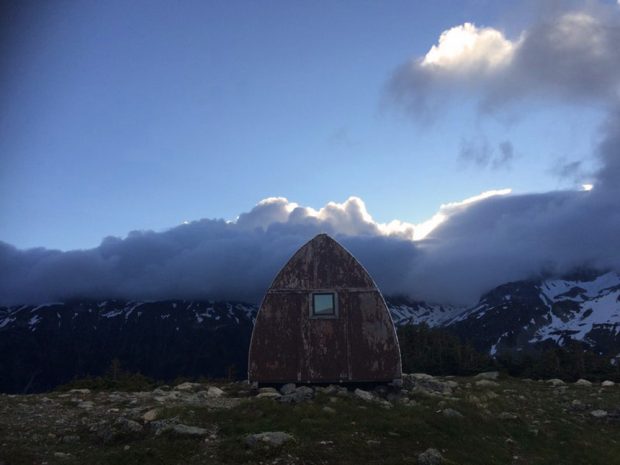 Le refuge à voûte de style gothique se dresse au milieu d’une prairie verdoyante ainsi que des blocs de pierre instables. Des nuages gris cachent les sommets au loin.