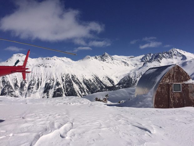 La queue et l’hélice du rotor de l’hélicoptère sont visibles à gauche de l’image et à droite se trouve le refuge Himmelsbach délabré, sous un ciel bleu pratiquement sans nuages.