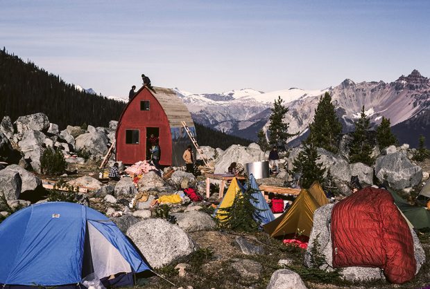 Les tentes et le matériel de camping se trouvent dans un pré rempli de galets devant le refuge voûté de style gothique. Les membres du club UBC-VOC s’emploient activement à la finition de la toiture et à s’occuper des autres aspects de la construction du refuge. En arrière-plan se dressent des pics enneigés.