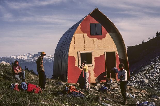 La charpente du refuge voûté de style gothique est achevée et les travaux de peinture ont commencé sur le mur d’extrémité avant. Les membres de l’équipe de bâtisseurs se tiennent devant le refuge et prennent un bain de soleil.