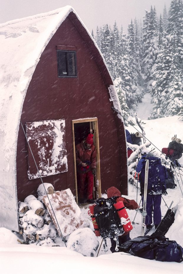Un skieur sort de l’entrée principale du refuge et deux autres se tiennent juste devant portant leur équipement de ski. La neige tombe et la paroi d’extrémité conserve sa couleur marron en dépit de la chute de neige blanche.