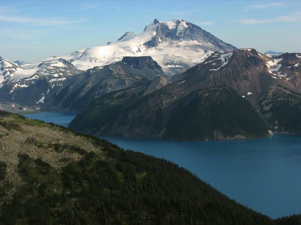 Le vent fait scintiller la couleur bleu profond du lac Garibaldi et les glaciers tout en haut miroitent au soleil. Le rocher brun et gris du pic montagneux dans l’arrière-plan se distingue des pentes jonchées d’arbres à feuilles persistantes d’un vert profond qui bordent le lac.