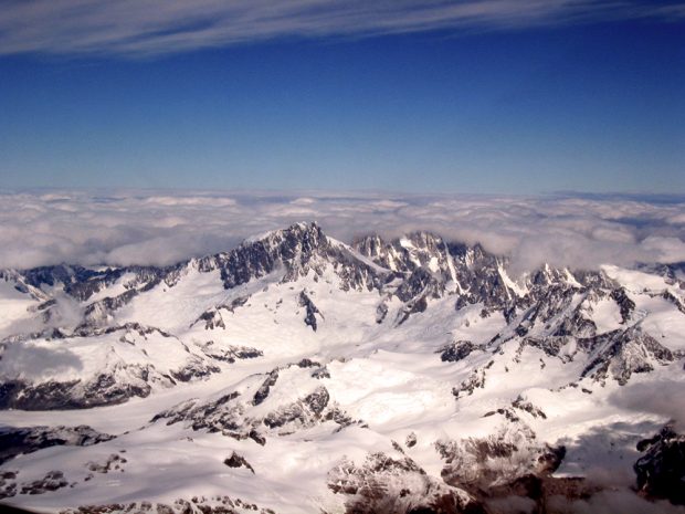 Le ciel bleu s’étend à distance au-dessus d’un champ glaciaire menant au pied du mont Waddington et le piton rocheux miroite au soleil à découvert; les autres pics à proximité du mont Waddington sont recouverts d’une bande de nuages bas et gris d’aspect orageux.