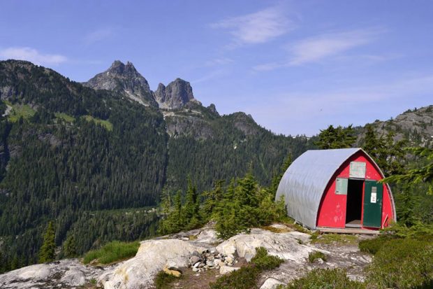 La photo a été prise à partir d’un angle et le refuge est situé à droite avec la porte principale grande ouverte. Les imposants pics se dressant en face de la vallée depuis le refuge se détachent des versants parsemés d’arbres à feuillage persistant.