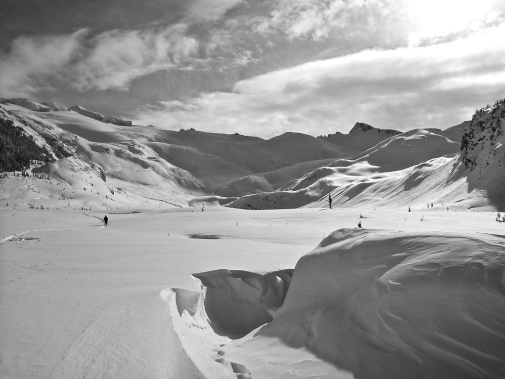 Une photo, noir et blanc, d’une vallée alpine recouverte de neige et des pics enneigés en arrière-plan. Une légère dépression indique l’emplacement du refuge et un skieur solitaire est aperçu au loin.