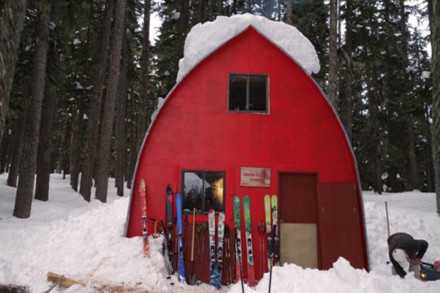 L’entrée du refuge a été déblayée, mais une grande plaque de neige est encore visible sur le toit. Des skis et des bâtons de skis sont adossés contre le mur d’extrémité avant. Un homme courbé en chapeau blanc déblaie l’entrée.