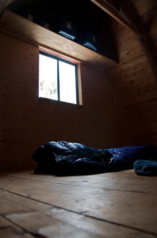 La lumière passe à travers la fenêtre et un grand sac de couchage bleu foncé est posé sur le plancher de bois. De grandes poubelles en plastique sont posées sur une grande étagère située au-dessus de la fenêtre.