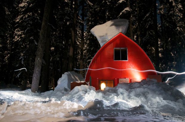 La cabane est entourée de neige pendant la nuit et les lumières de l’intérieur sont visibles à travers la fenêtre du rez-de-chaussée. Sur le toit voûté repose un petit amoncellement de neige.
