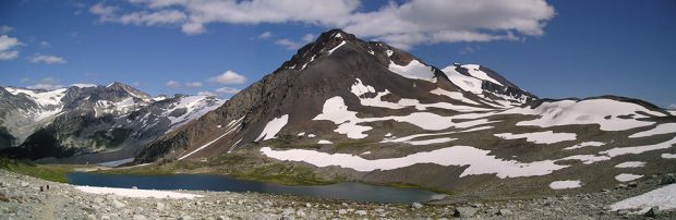 Un petit lac bleu entouré d’une petite pelouse en forme de cercle et un grand pic volcanique gris se dressant au-dessus avec des bancs de neige tout autour.