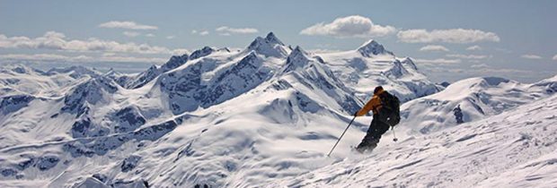 Un skieur en veste orange et portant un sac à dos noir prend le chemin opposé à celui du photographe sur une pente escarpée et enneigée, des pics recouverts de neige sont visibles au loin.