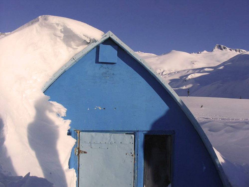 Mur avant bleu du refuge voûté de style gothique partiellement enfoui sous la neige, amoncelé d’un côté et dénudé de l’autre. Hauts versants enneigés et pics montagneux visibles en arrière-plan.