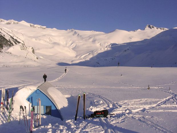 Le refuge a partiellement été déneigé; les skis et les bâtons de ski sont coincés dans la neige devant la cabane. Au loin, l’on aperçoit des skieurs se dirigeant vers le refuge et le soleil illumine les versants escarpés et enneigés en arrière-plan.