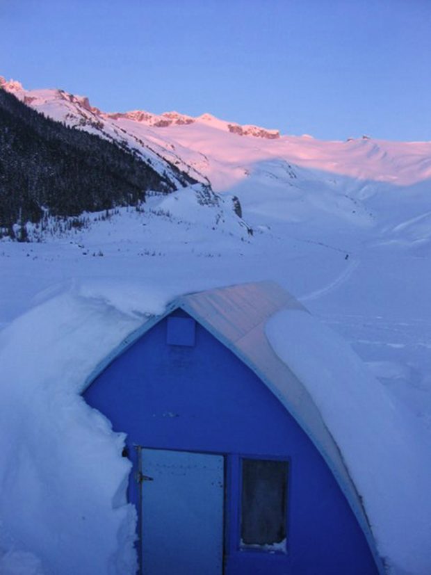 Le mur d’extrémité bleu, la porte principale et la fenêtre latérale ne sont pas recouverts de neige et le soleil couchant laisse une teinte rose-violacé sur les versants enneigés visibles au loin.