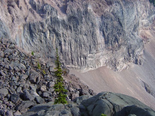 La paroi rocheuse rouge et grise à découvert illustre l’impact de l’érosion glaciaire. Au-dessous de la paroi, on aperçoit des amas de rochers érodés et arénacés ainsi qu’une coulée de blocs avec quelques arbustes à feuilles persistantes.