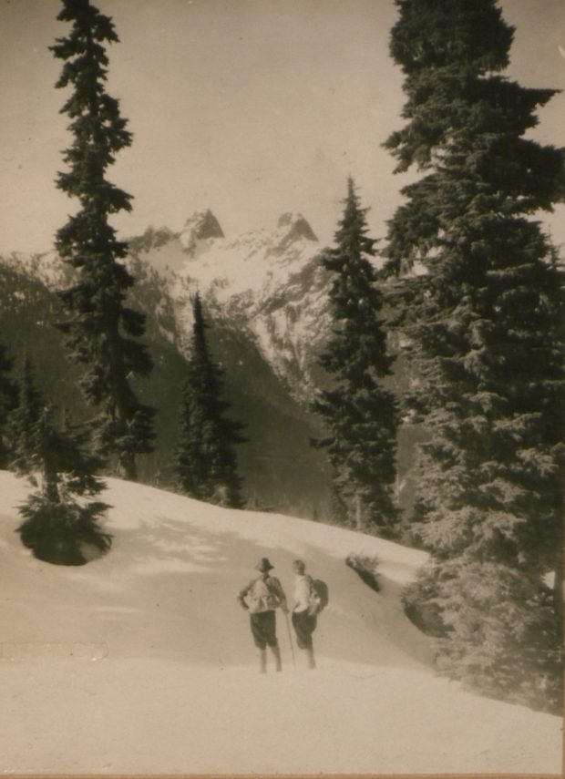 Un couple se tenant sur les pentes enneigées des montagnes du North Shore près de grands arbres à feuilles persistantes, le regard détourné de la caméra, admire les deux pics enneigés derrière appelés les Lions.