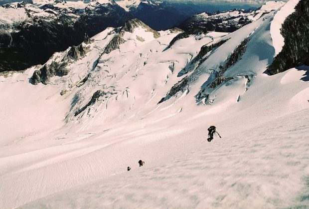 La perspective plongeante de l’image illustre une forte pente glaciaire et le premier de cordée portant un bonnet blanc et un sac à dos noir a presqu’atteint le sommet de la pente, l’on voit sa main toucher le sol enneigé. Les deux autres alpinistes aperçus plus bas le suivent.