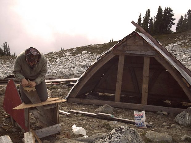 La pièce résultant de la toiture voûtée du refuge Brew 2 gît sur le sol. Au premier plan, un homme s’emploie activement au sciage d’un morceau de bois.