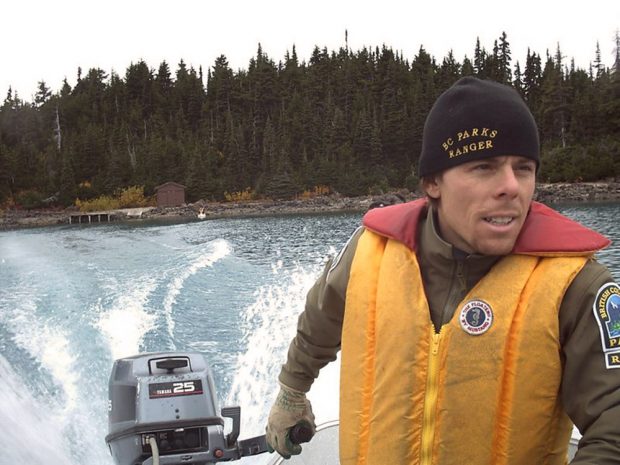 Portrait d’un homme en gilet de sauvetage jaune et rouge, tenant le gouvernail d’un moteur hors-bord. L’eau tourbillonne dans le sillage du bateau et au loin, on aperçoit le rivage à trois bandes.