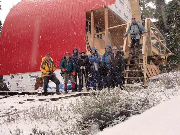 Portrait de groupe de l’équipe de bâtisseurs ayant aidé à l’érection du refuge par temps de chute de neige et avec des planches adossées contre le mur d’extrémité derrière eux sur un grand porche.