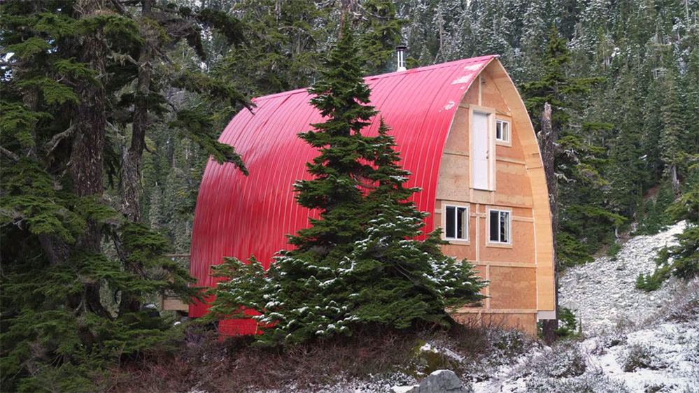 Le parement en aluminium rouge brillant recouvrant le toit du refuge en arc se démarque des arbres à feuilles persistantes vertes observés près du refuge. Une mince couche de neige recouvre le sol et les arbres près du refuge.
