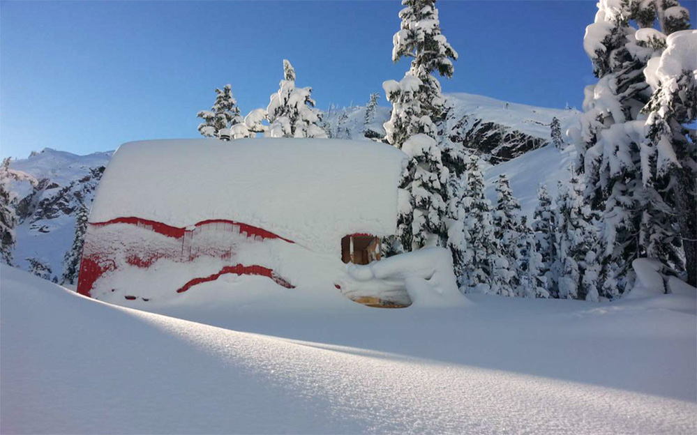 Une neige profonde a englouti le refuge et une partie du parement en aluminium rouge est à découvert. Le ciel hivernal arbore une couleur bleu clair et des arbres à feuillage persistant enneigés apparaissent en arrière-plan. 