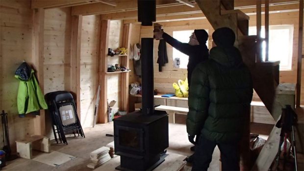 Un groupe de travailleurs s’emploie à l’installation de la cheminée noire de la poêle à bois. Des rayons de soleil traversent les fenêtres et les équipements sont empilés sur des étagères temporaires en arrière-plan.