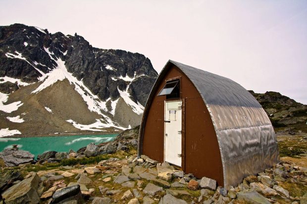 Entrée principale du refuge voûté de style gothique avec sa paroi d’extrémité brune et sa porte blanc brillant, près de l’eau turquoise du lac glaciaire. En arrière-plan, surgit un sommet sombre recouvert de couches de neige éparses.