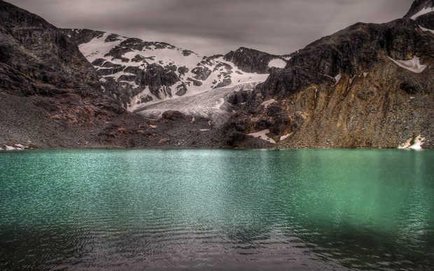 Une lueur du soleil se reflète sur l’eau turquoise. Le plan arrière illustre des berges rocheuses brun foncé et la partie supérieure du glacier s’érige en direction du piton rocheux nu et de nuages gris.
