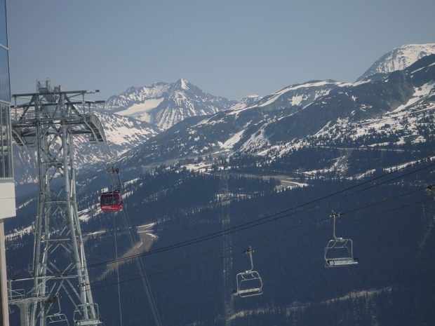 Une télécabine rouge descend d’un grand pylône d’acier sur Whistler Mountain et de l‘autre côté de la vallée on aperçoit le sommet de Blackcomb Mountain et d’Armchair Glacier.