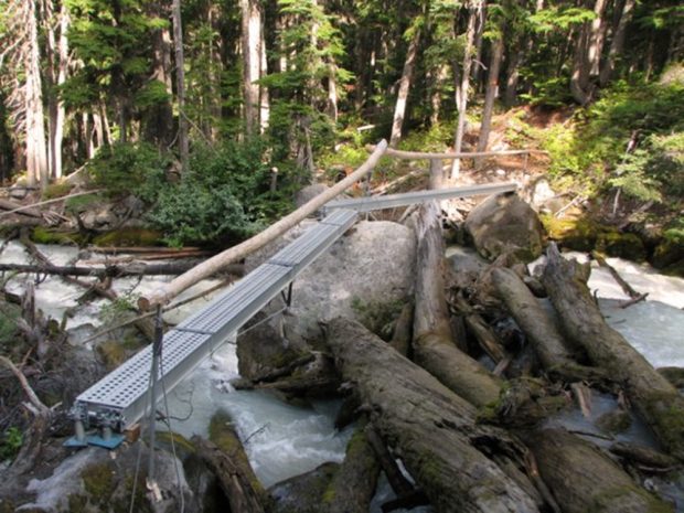 Passerelle en acier dotée d’une main courante en bois surplombant une rivière s’écoulant sur le sentier du refuge Harrison avec une forêt à feuillage persistant visible au loin sur la rive du ruisseau.