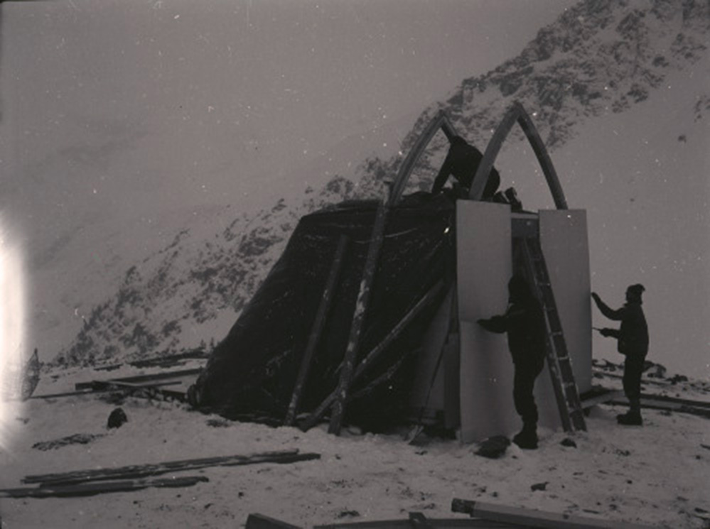 La neige couvre le sol de même que le versant et trois membres de l’équipe de construction essaient d’assembler le refuge.