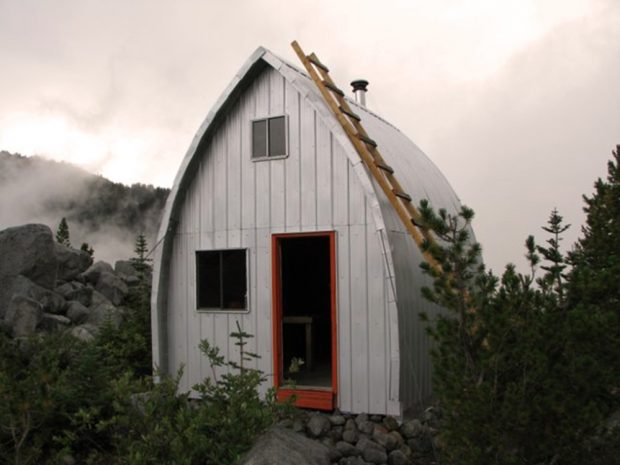 Le refuge Harrison entièrement paré d’un revêtement en aluminium après sa rénovation en août 2014. Des nuages blancs tourbillonnent dans le ciel couvert et des arbres à feuilles persistantes sont visibles en face de la structure argentée et brillante du refuge rénové.