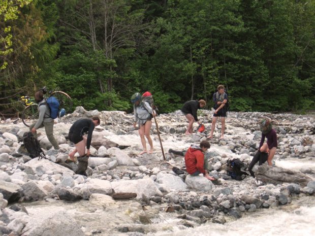 Un groupe de membres de l’UBC-VOC profitant du courant de la crique pour se baigner après avoir travaillé à l’aménagement du sentier menant au refuge Harrison. L’un des membres porte une bicyclette et des sacs pour traverser la crique s’écoulant.