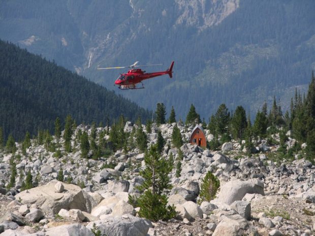 Un hélicoptère de couleur rouge vif survole le refuge Harrison avant sa rénovation. La façade d’extrémité orange est visible dans la coulée de pierres l’entourant. Des arbustes à feuilles persistantes vertes poussent près du refuge.