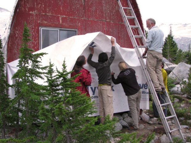 Tyvek, une membrane en plastique blanche, appliquée à la façade d’extrémité altérée par les intempéries. Roland Burton se tenant sur une échelle argentée, le regard tourné vers 4 membres tenant la membrane Tyvek.