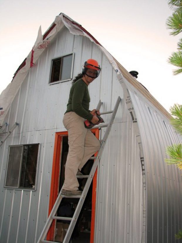 Portrait d’un homme portant un casque orange et tenant une perceuse électrique, debout sur une échelle en aluminium se trouvant près de l’entrée principale du refuge.