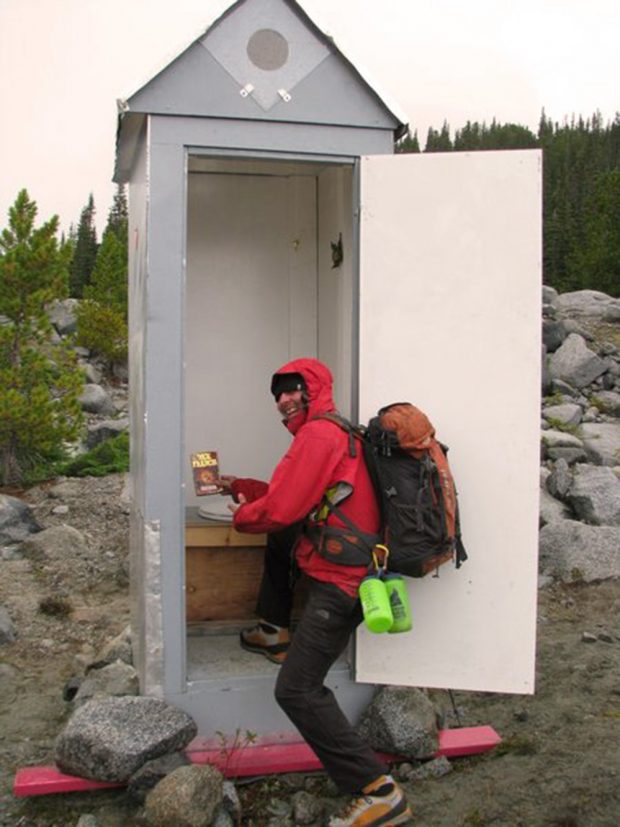 L’image montre un homme tenant dans sa main droite un livre de poche, vêtu d’un blouson à capuche rouge et portant un sac à dos gris avec un pied à l’intérieur des toilettes extérieures nouvellement aménagées.