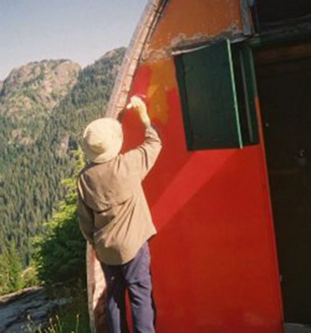 Un homme coiffé d’un chapeau blanc repeint le mur d’extrémité avant orange défraîchi du refuge en rouge vif.