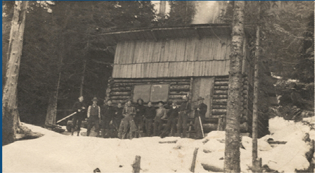 Photographie en noir et blanc d'un groupe d’hommes faisant la queue devant la cabane en rondins. La neige couvre le sol autour de la cabane et les arbres en arrière-plan sont dépouillés.