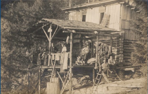 Photographie en noir et blanc de la cabane; les membres posent sur les escaliers, accoudés à la balustrade sous le porche. Cette dernière est drapée d’un linge.