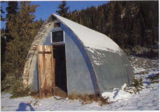 Vue de l’entrée principale d’un refuge à voûte de style gothique dont la porte est entrebâillée. Une mince couche de neige est visible sur le toit en voûte, et des arbres à feuilles persistantes apparaissent en arrière-plan.