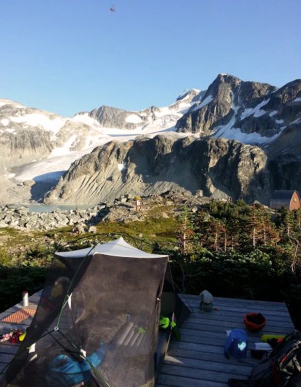 Une tente-filet noire se dresse sur une plateforme en bois, le refuge se trouve dans la prairie verte parsemée d’arbres et, en arrière-plan, le sommet du mont Wedge.