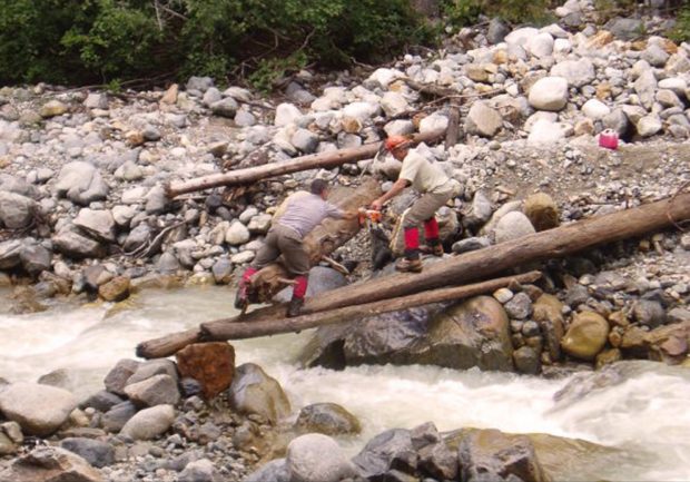 Deux hommes se tenant sur un arbre qui a été hissé en travers d’un cours d’eau au débit rapide essaient de déplacer une autre grosse bille de bois à côté de celle sur laquelle ils se tiennent.