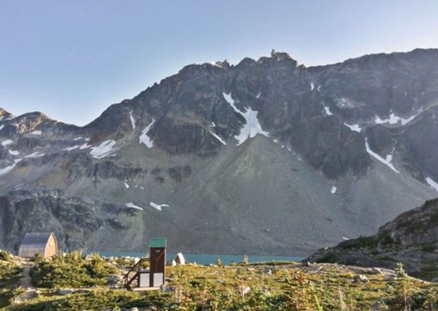 Les bécosses au toit vert peintes en marron se dressent sur la prairie parsemée d’arbustes et de petits rochers; le refuge se trouve à gauche. Derrière se trouve l’eau turquoise du lac et le roc gris foncé de la cime de la montagne.